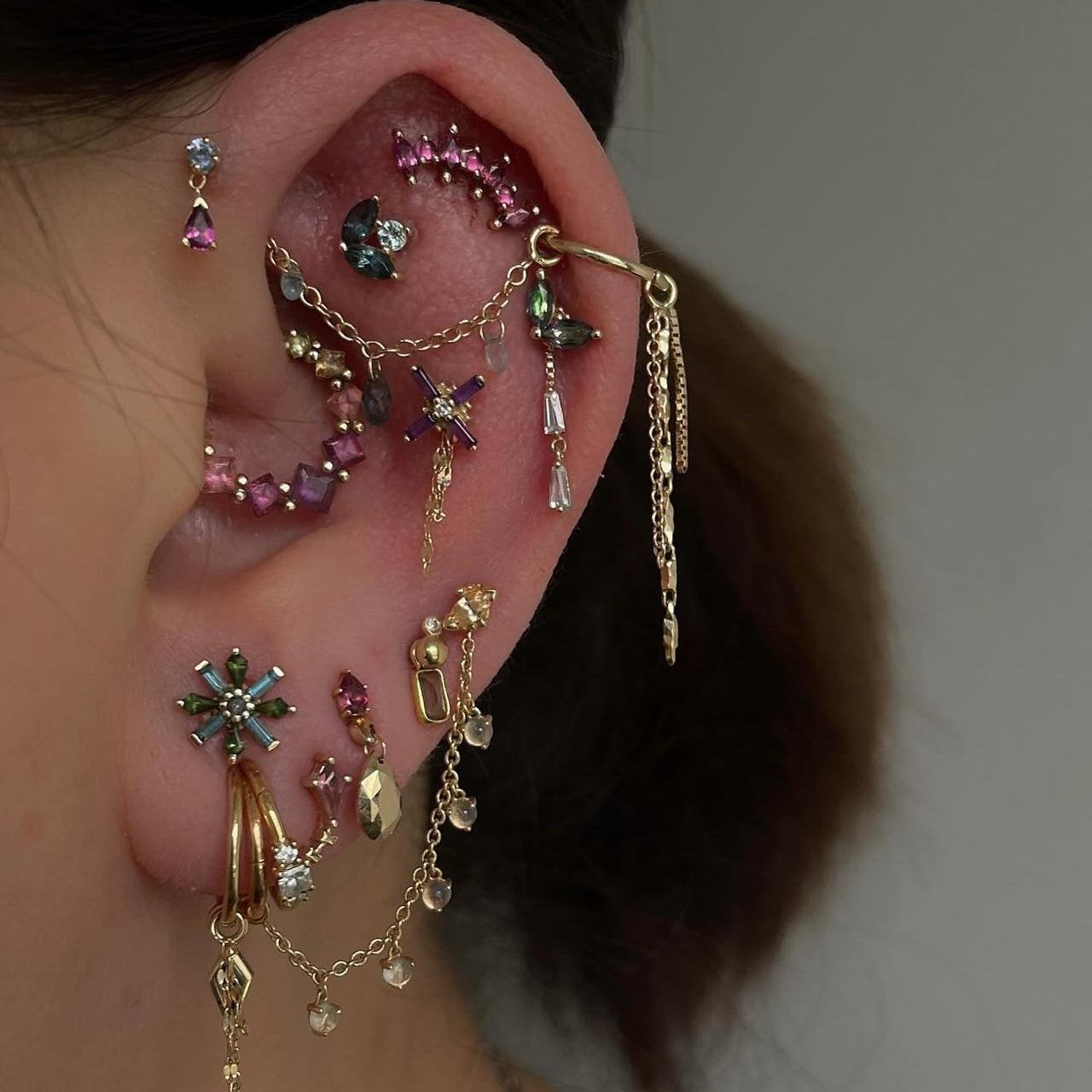 Close-up of an ear with multiple gold and gemstone earrings against a neutral background